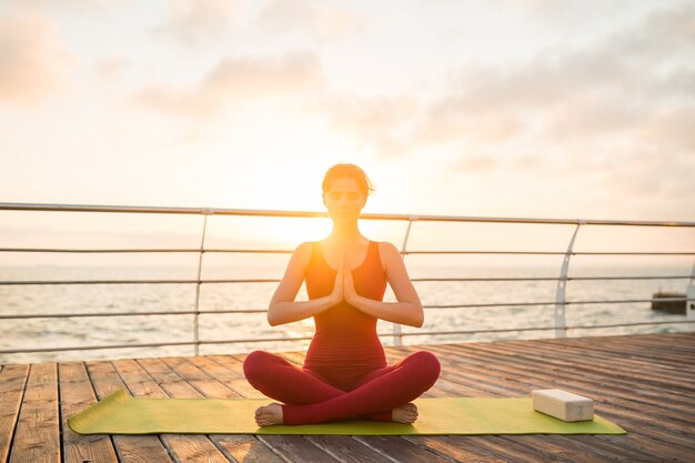 Joven mujer atractiva hermosa delgada haciendo yoga por la mañana al amanecer por el mar, estilo de vida saludable