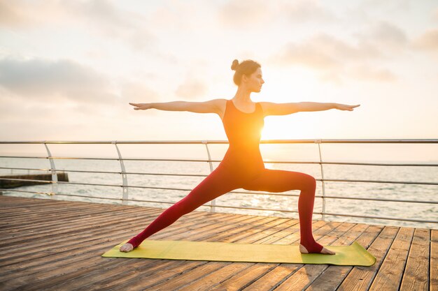 Joven mujer atractiva hermosa delgada haciendo yoga por la mañana al amanecer por el mar, estilo de vida saludable