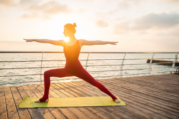 Joven mujer atractiva hermosa delgada haciendo yoga por la mañana al amanecer por el mar, estilo de vida saludable