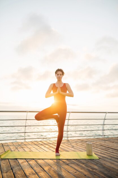 Joven mujer atractiva hermosa delgada haciendo yoga por la mañana al amanecer por el mar, estilo de vida saludable