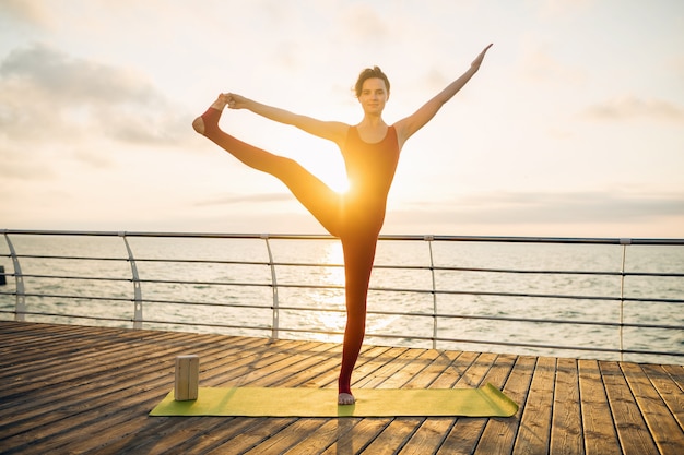 Joven mujer atractiva hermosa delgada haciendo yoga por la mañana al amanecer por el mar, estilo de vida saludable