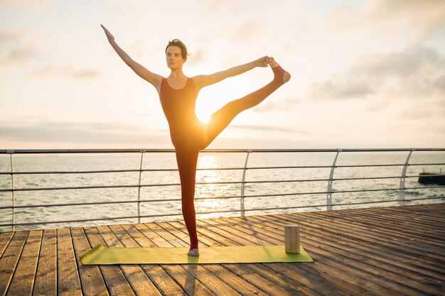 Joven mujer atractiva hermosa delgada haciendo yoga por la mañana al amanecer por el mar, estilo de vida saludable