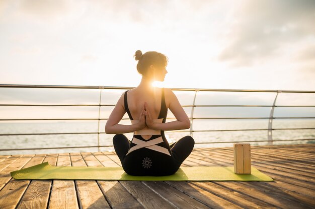 Joven mujer atractiva hermosa delgada haciendo yoga por la mañana al amanecer por el mar, estilo de vida saludable