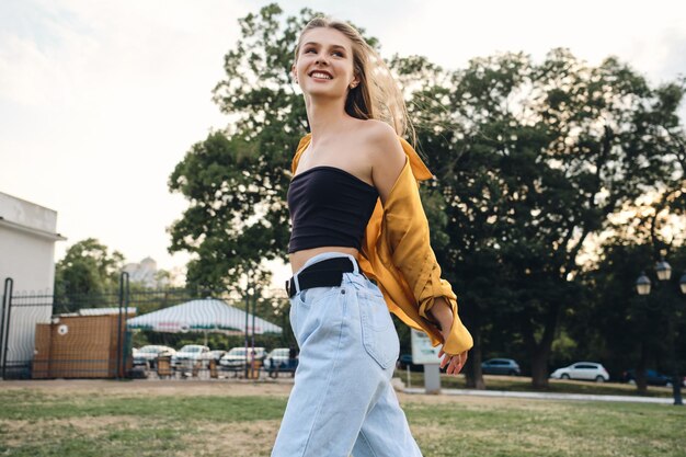 Joven mujer bastante alegre con camisa amarilla y jeans caminando felizmente sobre el césped en el parque de la ciudad
