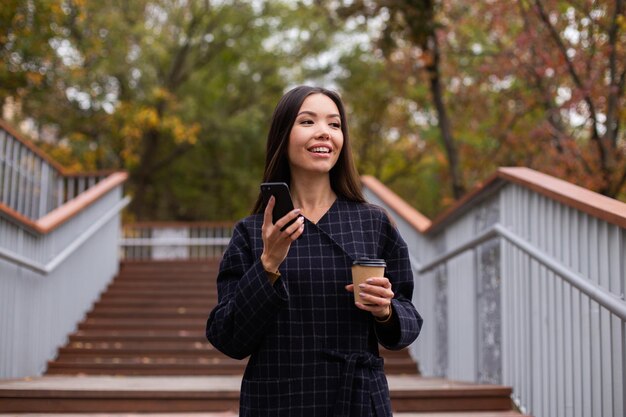 Joven mujer bastante casual en abrigo con café para llevar y teléfono celular felizmente mirando hacia otro lado en el parque de la ciudad