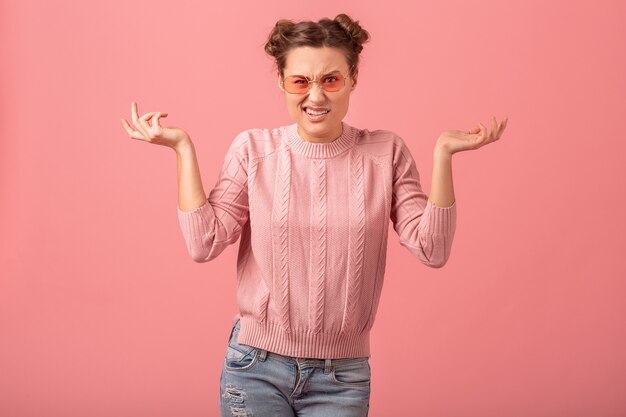 Joven mujer bastante divertida que tiene un problema, siente estrés, levanta las manos en un suéter rosa y gafas de sol aisladas sobre fondo rosa de estudio