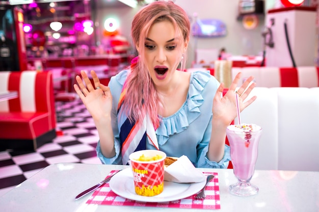 Joven mujer bonita y encantadora con colores de cabello rosados inusuales, sentada en un café americano vintage, expresivas emociones positivas de sorpresa, ropa y accesorios vintage pastel, disfruta de su comida rápida