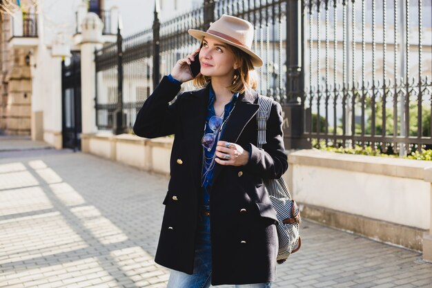 Joven mujer bonita con estilo sonriendo y hablando por su teléfono, vestida con abrigo azul oscuro y jeans