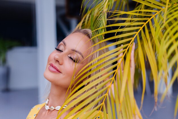 Joven mujer bonita feliz tranquila caucásica con maquillaje de noche brillante con vestido amarillo de verano y collar de conchas de mar