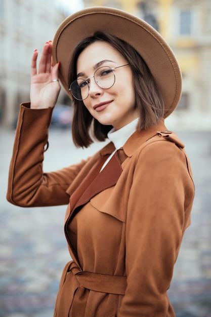 Joven mujer bonita en gafas modernas y sombrero de moda y abrigo marrón está posando en el centro de la ciudad