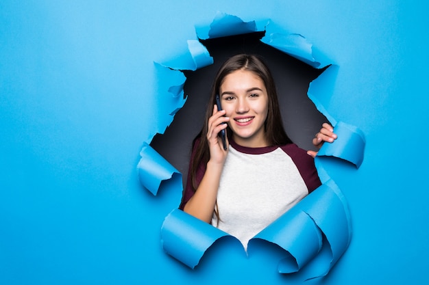 Joven mujer bonita hablar teléfono mientras mira a través del agujero azul en la pared de papel.