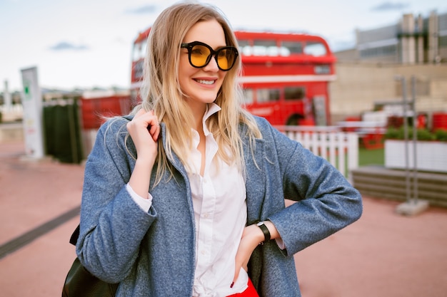 Joven mujer bonita rubia caminando en el centro de la ciudad de Londres, vistiendo un elegante atuendo de estudiante informal, abrigo azul y gafas de colores, otoño primavera a mitad de temporada, humor de viaje.