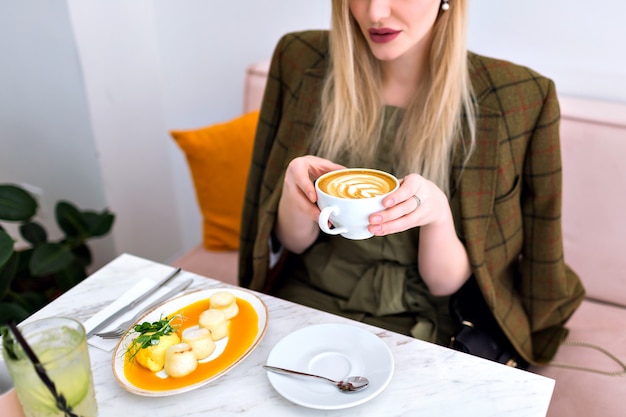 Joven mujer bonita rubia disfrutando de un sabroso brunch saludable con tostadas de salmón y aguacate, capuchino, limonada y postre, traje elegante, interior elegante y ligero, sosteniendo una taza de café.