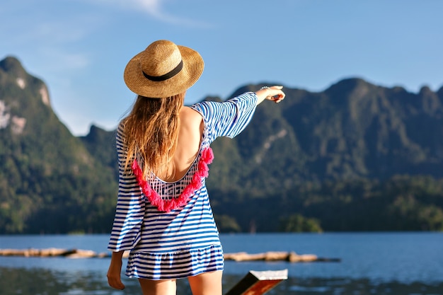 Joven mujer bonita rubia con pelos largos, con sombrero vintage y un vestido de moda sexy brillante mira hacia las montañas y el lago, muestra su mano, increíbles aventuras de verano.