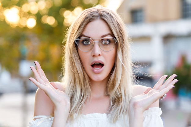 Joven mujer bonita rubia sonriendo y haciendo cara linda, gafas transparentes y camisa blanca, posando en el parque de la ciudad