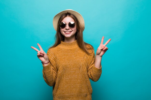 Joven mujer bonita con sombrero y gafas de sol señaló gesto de paz en la pared azul