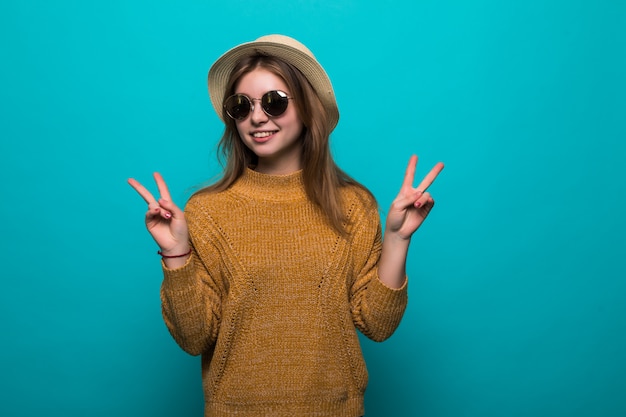 Joven mujer bonita con sombrero y gafas de sol señaló gesto de paz en la pared azul