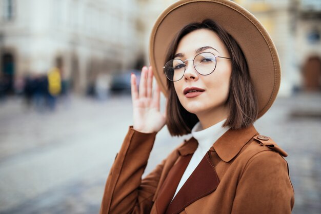 Joven mujer bonita con sombrero de moda y abrigo marrón está posando en el centro de la ciudad