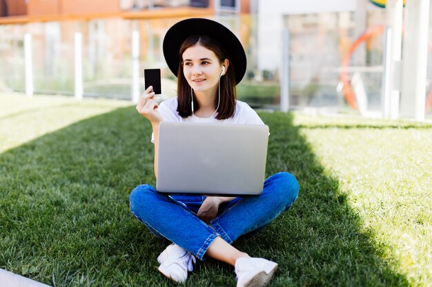 Joven mujer bonita con tarjeta de crédito y usando la computadora portátil para la compra mientras está sentado en la hierba verde en el parque
