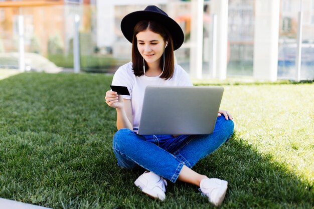 Joven mujer bonita con tarjeta de crédito y usando la computadora portátil para la compra mientras está sentado en la hierba verde en el parque