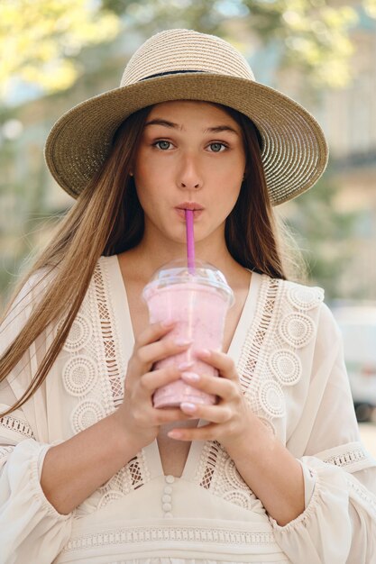 Joven mujer bonita con vestido blanco y sombrero bebiendo batidos mirando soñadoramente a la cámara mientras estaba sola en la calle de la ciudad