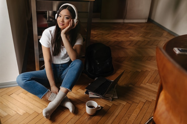 Joven mujer bronceada en jeans con estilo y calcetines blancos está mirando al frente y posando en auriculares