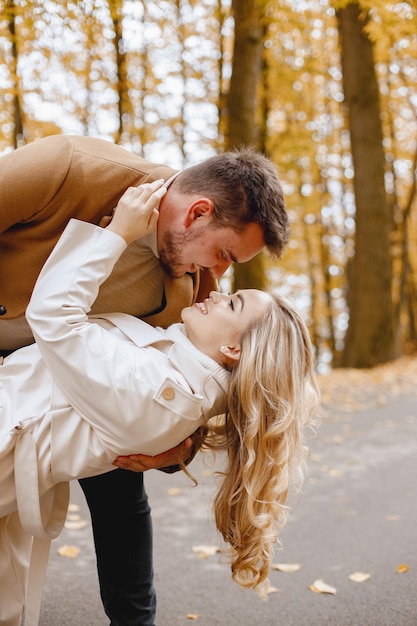 Joven y mujer caminando afuera con abrigos beige. Mujer rubia y hombre moreno en el bosque de otoño. Pareja romántica besándose.