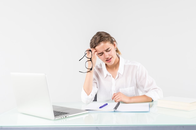 Joven mujer cansada delante de una computadora portátil en el escritorio de oficina, aislado sobre fondo blanco.
