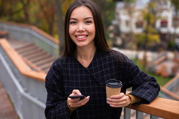 Joven mujer casual alegre en abrigo con café para llevar y teléfono celular felizmente mirando en cámara en el parque de la ciudad