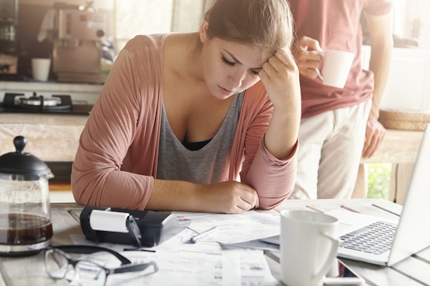 Joven mujer casual con aspecto deprimido mientras administra las finanzas familiares y hace papeleo, sentado en la mesa de la cocina con muchos papeles, calculadora y computadora portátil