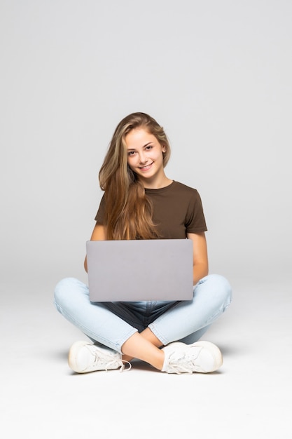 Joven mujer casual sentada sonriendo sosteniendo portátil aislado en la pared blanca