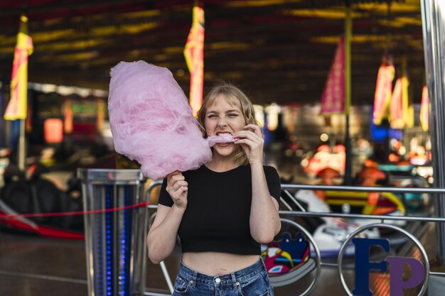 Joven mujer caucásica alegre comiendo algodón de azúcar en un parque de atracciones