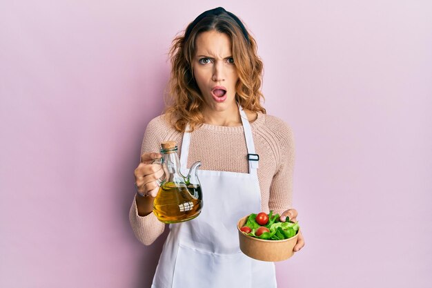 Joven mujer caucásica con delantal sosteniendo una lata de aceite de oliva y ensalada con cara de sorpresa, con aspecto escéptico y sarcástico, sorprendida con la boca abierta