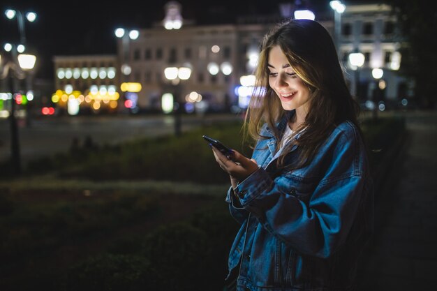 Joven mujer caucásica enviando mensajes de texto por teléfono celular en la ciudad por la noche