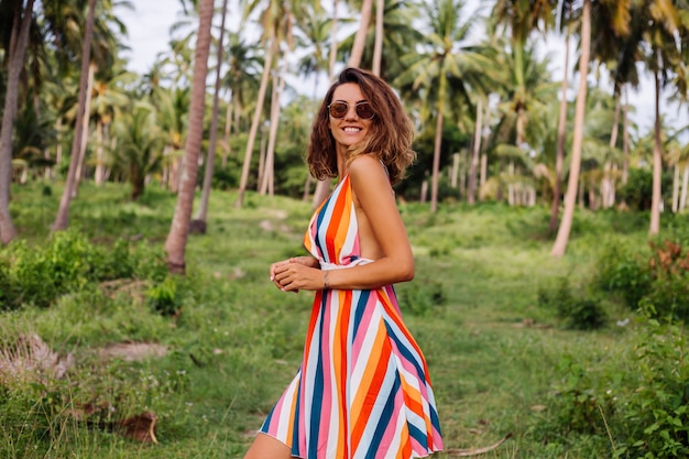 Joven mujer caucásica feliz en vestido de verano a rayas de colores con pelo corto y rizado en gafas de sol Vacaciones en un país cálido y exótico.