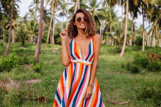 Joven mujer caucásica feliz en vestido de verano a rayas de colores con pelo corto y rizado en gafas de sol Vacaciones en un país cálido y exótico.