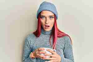 Foto gratuita joven mujer caucásica con gorra de invierno y bebiendo una taza de café caliente con cara de shock que parece escéptica y sarcástica sorprendida con la boca abierta