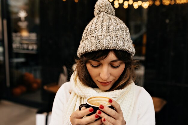 Joven mujer caucásica en gorro de punto tomando café y caminando por las calles con luces de humor navideño