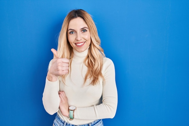 Joven mujer caucásica de pie sobre fondo azul haciendo feliz gesto de aprobación con la mano. expresión de aprobación mirando a la cámara que muestra el éxito.