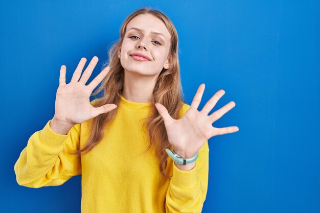 Joven mujer caucásica de pie sobre fondo azul que aparece y señala con los dedos número diez mientras sonríe confiada y feliz.