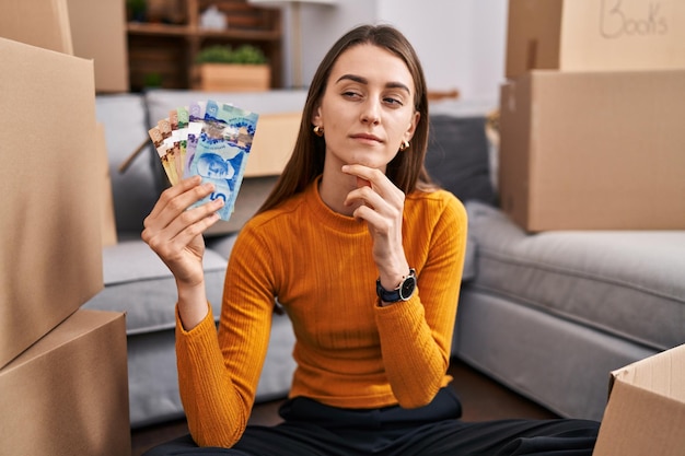 Joven mujer caucásica sentada en el suelo en una casa nueva con dinero canadiense cara seria pensando en la pregunta con la mano en la barbilla, pensativa sobre una idea confusa