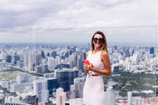 Joven mujer caucásica sonriente feliz viajero en vestido apropiado y gafas de sol en piso alto en bangkok sosteniendo el teléfono