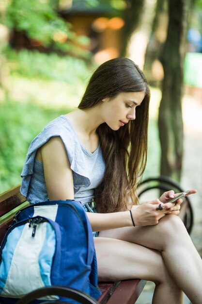 Joven mujer caucásica con un teléfono celular, sentada en un parque en un banco de madera, leyendo un SMS.