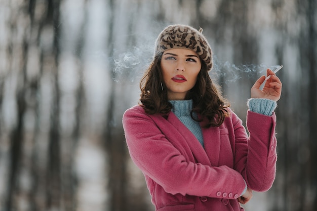 Joven mujer caucásica vistiendo un abrigo rosa y fumando en un parque con árboles en el fondo