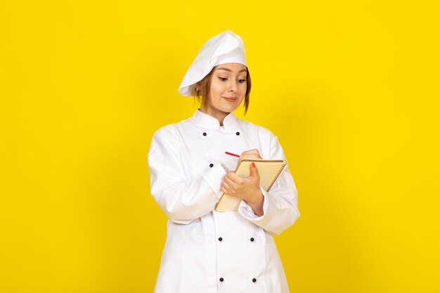 Joven mujer cocinando en traje de cocinero blanco y gorra blanca escribiendo notas