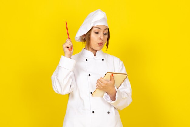 Joven mujer cocinando en traje de cocinero blanco y gorra blanca escribiendo notas