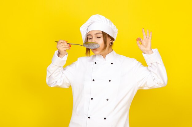 Joven mujer cocinando en traje de cocinero blanco y gorra blanca posando pensando sosteniendo una cuchara de plata degustando