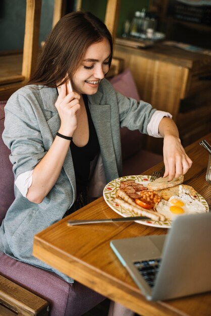 Joven mujer comiendo comida en el restaurante hablando por teléfono móvil