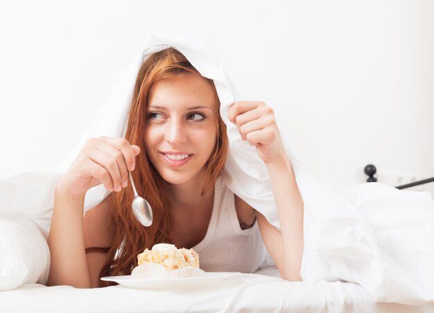 Joven mujer comiendo pastel dulce debajo de la sábana en la cama