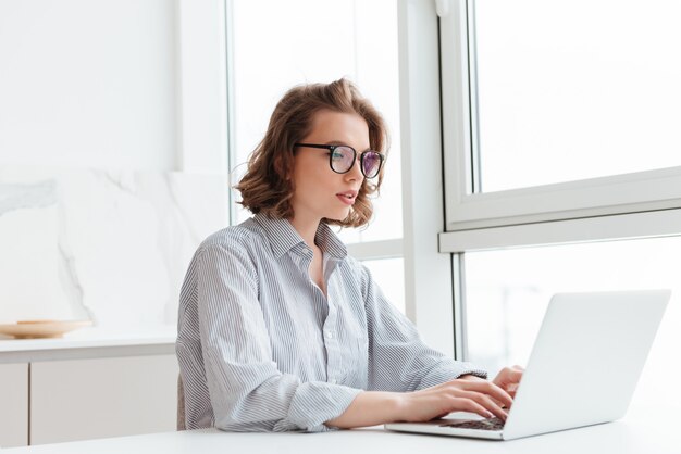 joven mujer concentrada en camisa a rayas usando laptop mientras se sienta a la mesa en el apartamento de luz
